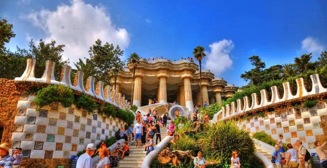 Muros y escaleras con mosaicos llevan a la Sala Hipóstila en el Park Güell, Barcelona, con numerosos turistas en un día solea
