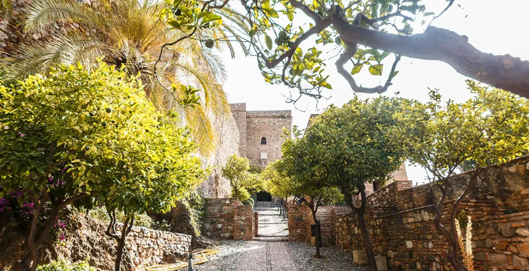 Kopfsteinpflasterweg führt zu einer alten Steinfestung in Malaga, gesäumt von Zitrus- und Palmenbäumen sowie Ziegelmauern.