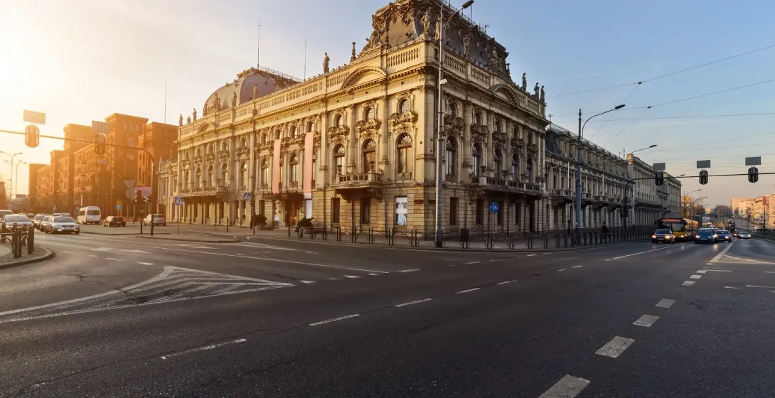 Zdobiony, jasny pałac w stylu neorenesansowym na słonecznym rogu miejskiej ulicy z sygnalizacją świetlną i samochodami.