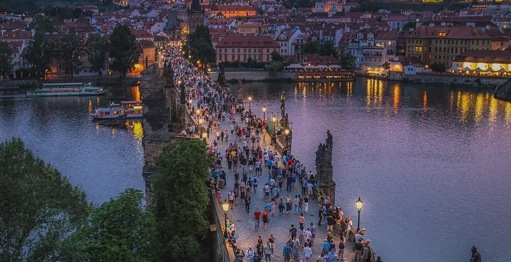 Tourists stroll across Charles Bridge in Prague at sunset, with Prague Castle majestically lit in the background.