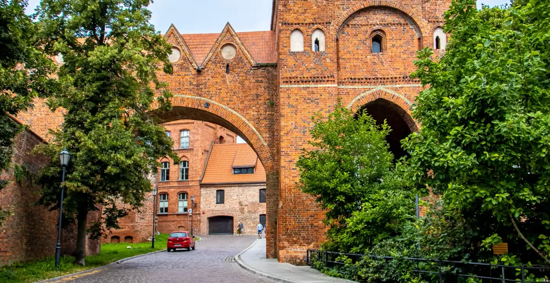 Brukowana ulica prowadząca do zabytkowej bramy miejskiej z czerwonym dachem, otoczonej zielonymi drzewami, żółty parasol na p