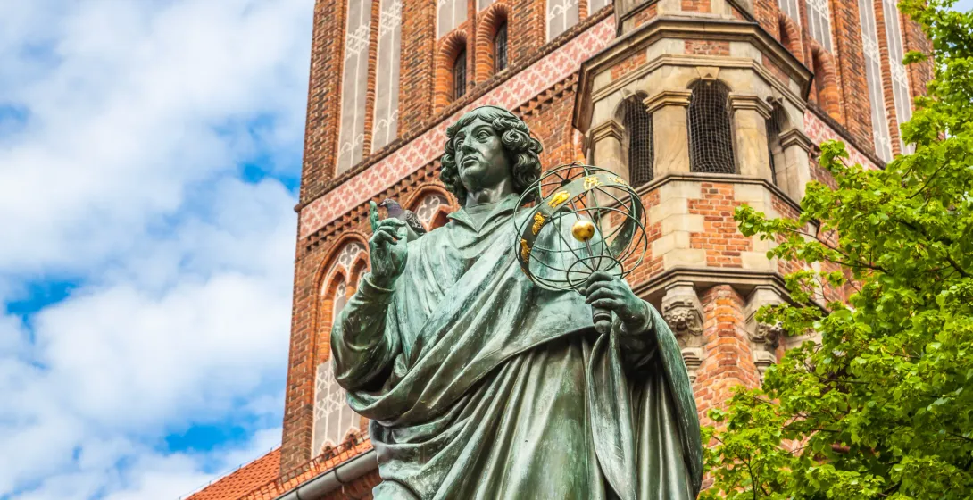Brązowy posąg Mikołaja Kopernika trzymającego sferę armilarną, z zabytkową ceglaną wieżą kościoła i błękitnym niebem w tle.