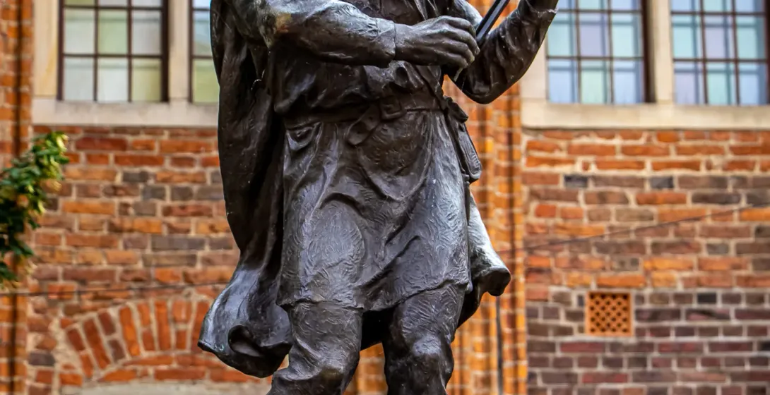 Brązowy posąg bosego skrzypka grającego na skrzypcach, stojący na białym cokole przed ceglanym budynkiem z oknami.