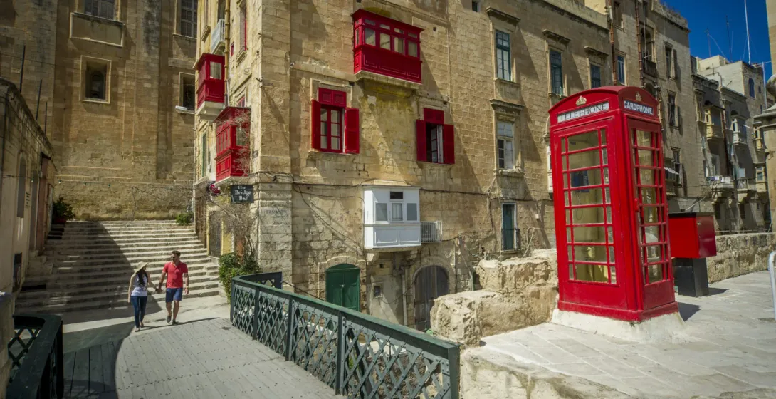 Pareja caminando por un puente en La Valeta, Malta, con edificios históricos y una cabina telefónica roja.