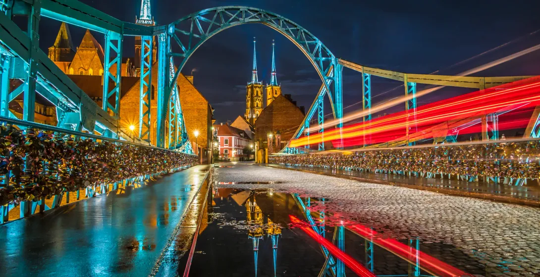Tumski Bridge in Wroclaw at night, illuminated in blue, with many love locks on railings and blurred red light trails. Ostrów