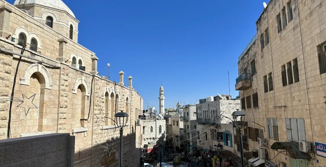 A narrow Bethlehem street with a church dome and cross on one side and a minaret in the distance.
