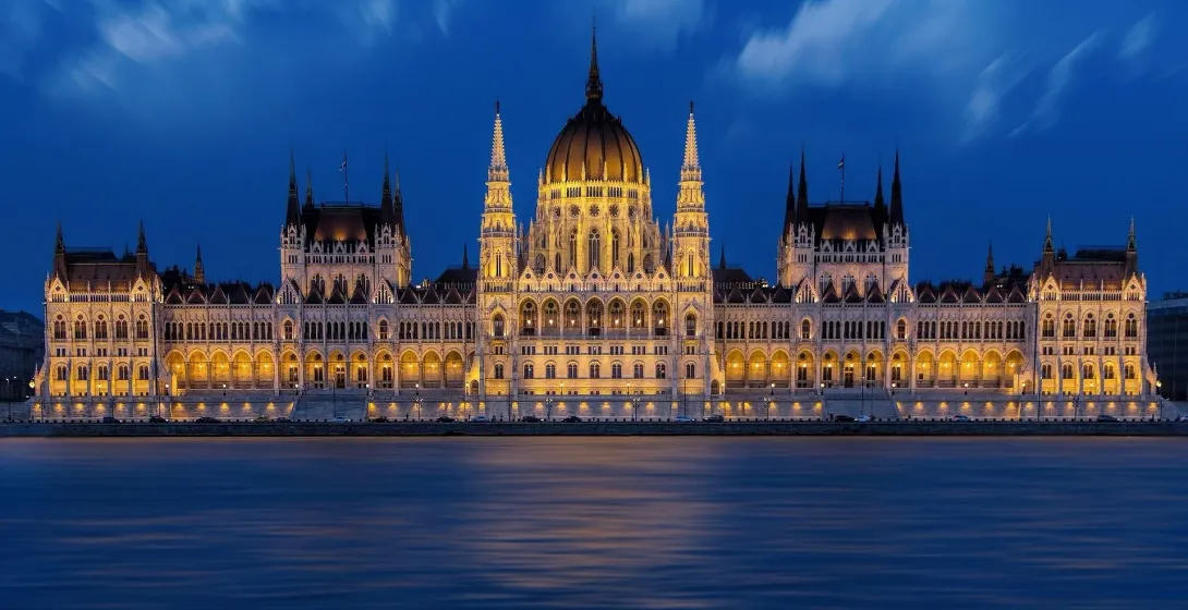 Illuminated Hungarian Parliament building at night, viewed from across the Danube River with blue sky.