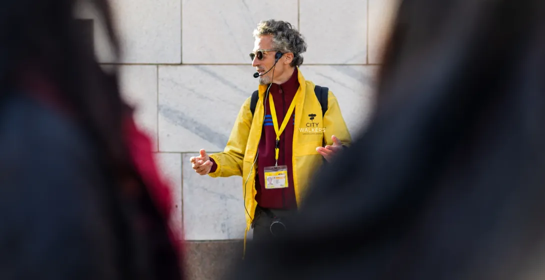 Tour guide in a yellow jacket and headset microphone speaks to a group outdoors.