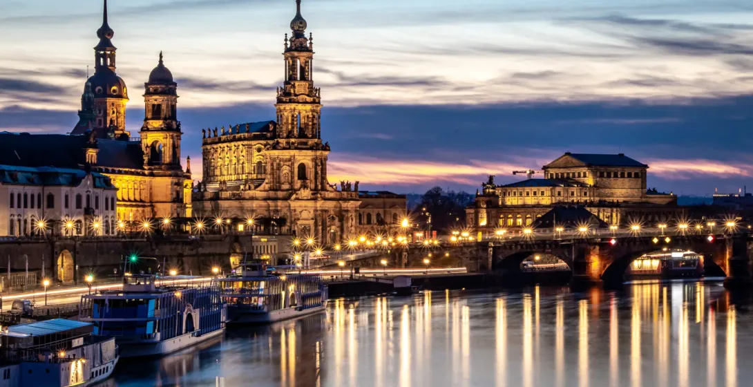 Dresden skyline at twilight over the Elbe River, with illuminated historic buildings and reflections on the water.