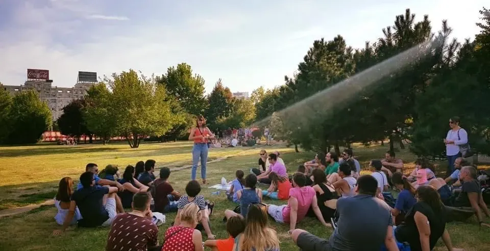 A tour guide speaks to a group of tourists seated on the grass in a sunny park. A large building with a Coca-Cola sign is ...