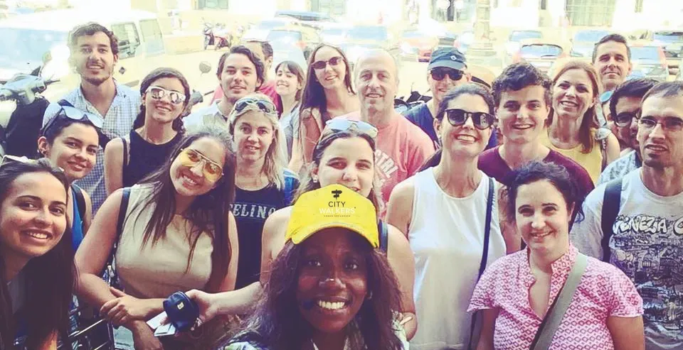 Tour guide in a yellow cap smiles for a group selfie with walking tour participants in a sunny city square.