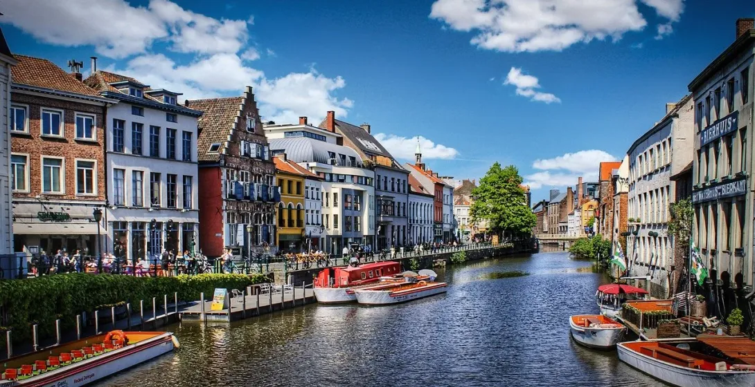 A sunny view of a canal in Ghent lined with historic, colorful buildings and several tour boats docked along the side.