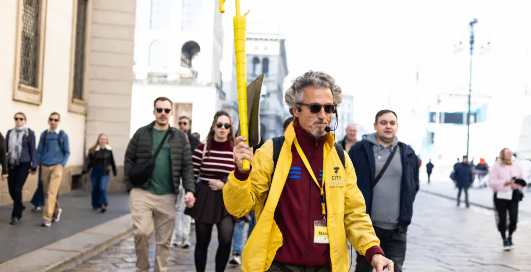 A tour guide in a yellow jacket holds up a yellow umbrella while leading a walking tour through a city street.