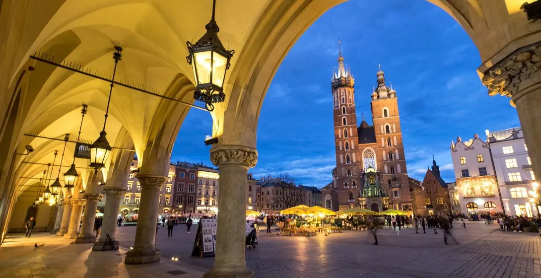 View from an illuminated archway of the Krakow Main Market Square with St. Mary's Basilica at dusk.