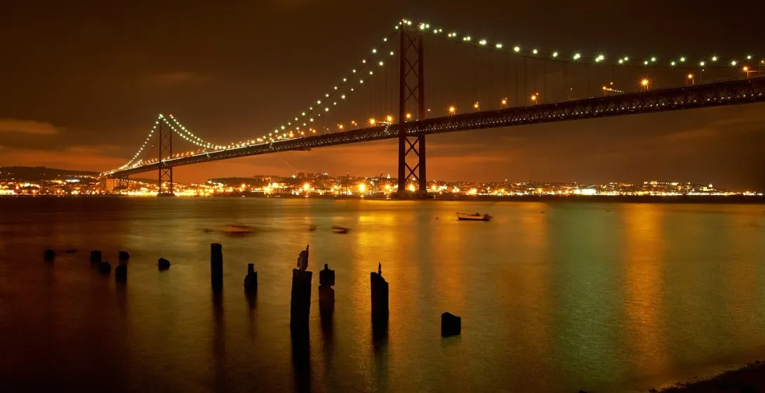 25 de Abril Bridge illuminated at night over the Tagus River, with city lights and reflections on the water.