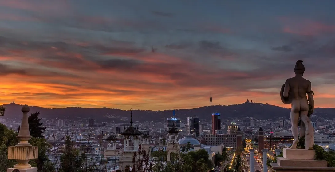 Widok na panoramę Barcelony o zachodzie słońca ze statuą na pierwszym planie i górą Tibidabo w tle.