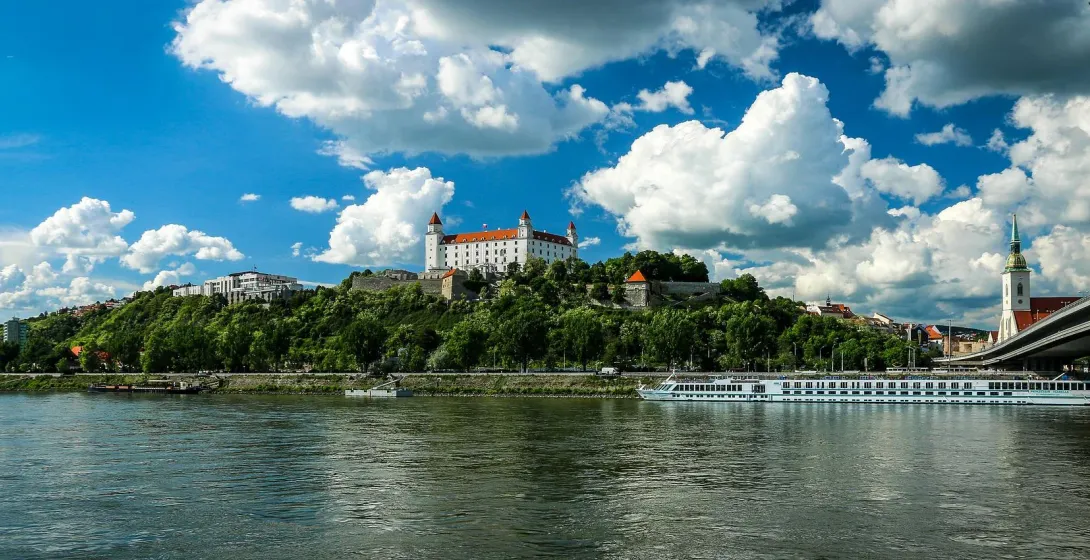 Bratislava Castle with red roofs on a green hill overlooking the Danube River with a boat, under a blue sky.