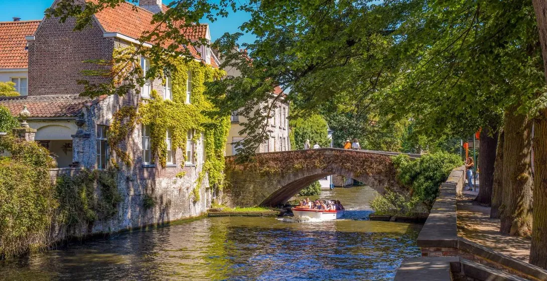 A canal boat with tourists passes under a historic cobblestone bridge in Bruges, bordered by ivy-covered buildings.