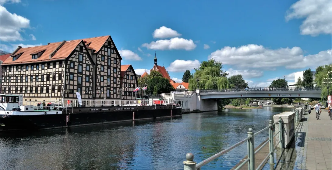 Half-timbered buildings with red roofs on the riverbank, a long barge, a concrete bridge, and a pedestrian path under a blue