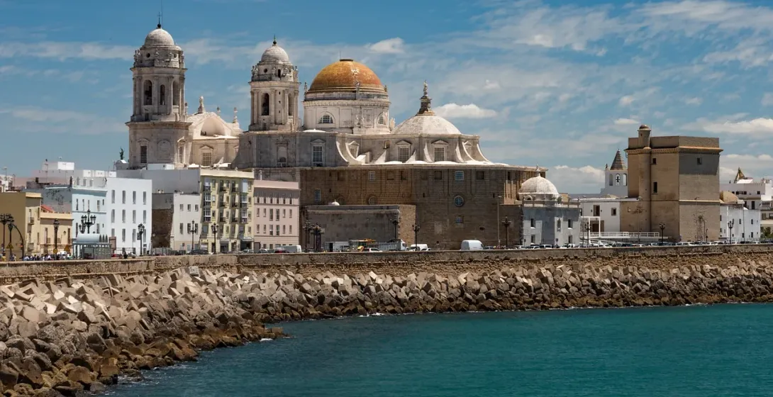 View of Cádiz Cathedral's domes and bell towers alongside other city buildings from across the turquoise sea.