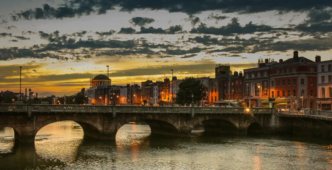 Dublin cityscape at sunset with Ha'penny Bridge over the River Liffey, glowing warm light, and historical buildings.
