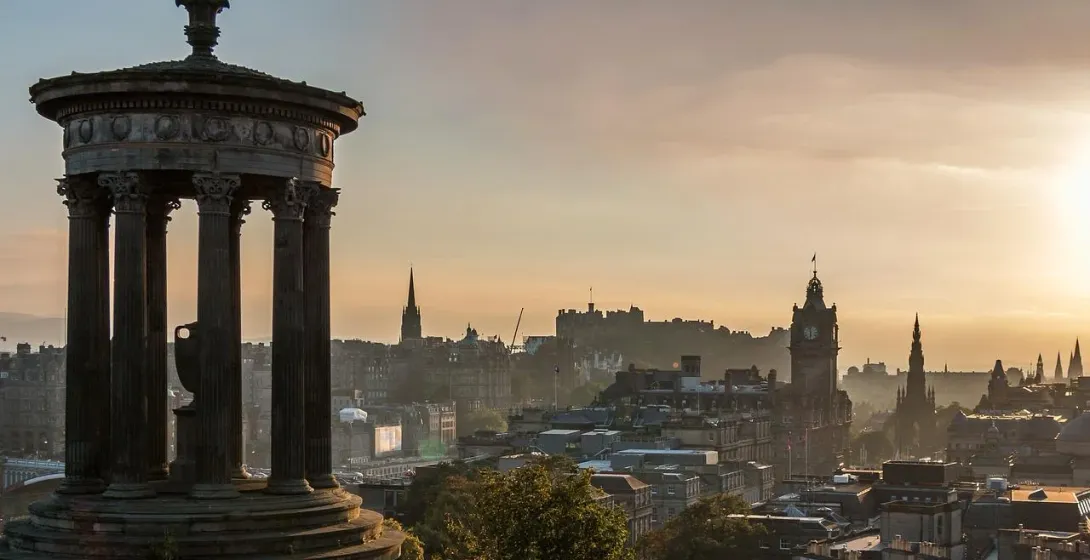 Dugald Stewart Monument foreground, Edinburgh Castle, and city skyline at sunrise with hazy sky.