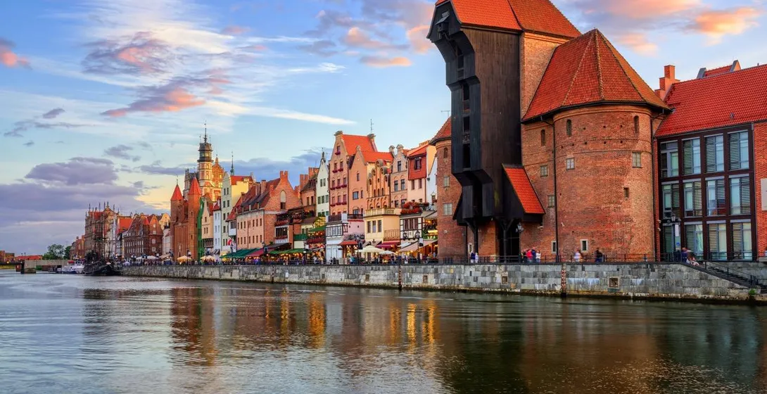 Historic waterfront buildings and a large wooden crane reflecting in the Motława River at sunset in Gdańsk.