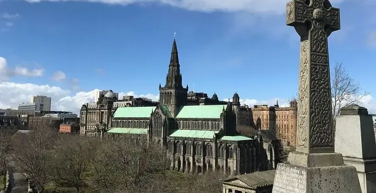 Glasgow Cathedral with green roof, seen from a historic cemetery with a large Celtic cross in the foreground.