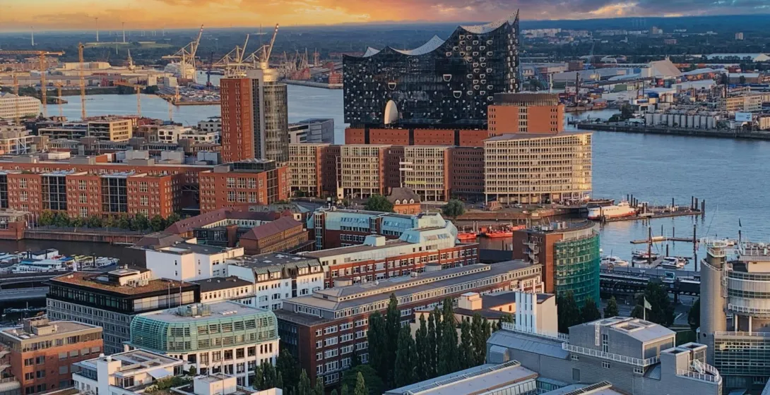 Aerial view of Hamburg harbor at sunset, featuring the Elbphilharmonie, brick buildings, and industrial cranes by the water.