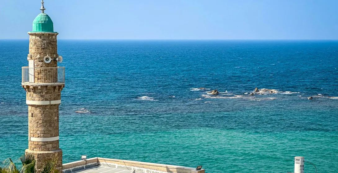 Minarete de piedra con cúpula verde y balcón, dominando el mar Mediterráneo turquesa, con tejados y follaje verde.