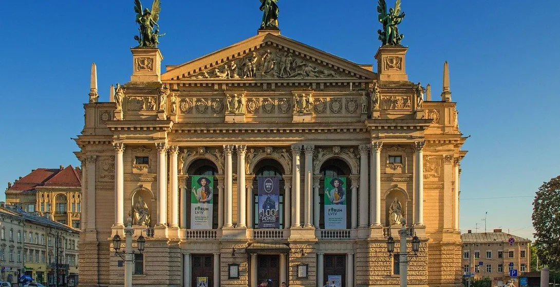 Fassade der Lwiwer Oper mit Säulen, Skulpturen auf dem Dach und Bannern. Klarer blauer Himmel. Menschen gehen im Vordergrund.