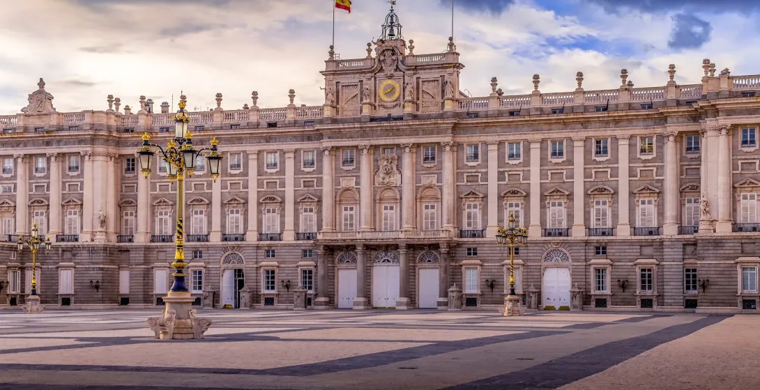 Exterior view of the Royal Palace of Madrid under a dramatic sky, with cobblestone plaza and lampposts.