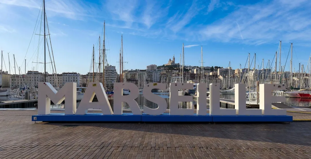 Große MARSEILLE-Buchstaben auf blauem Sockel am Vieux-Port, mit Segelbooten und Gebäuden. Notre-Dame de la Garde auf dem Hüge