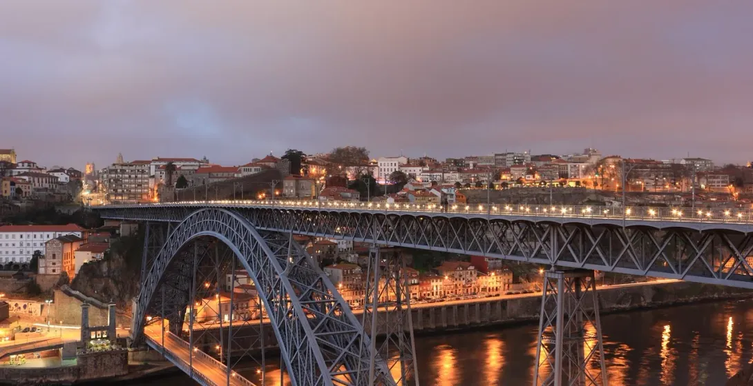 Large metal arch bridge with two decks illuminated at dusk over a river, with city buildings on both sides.