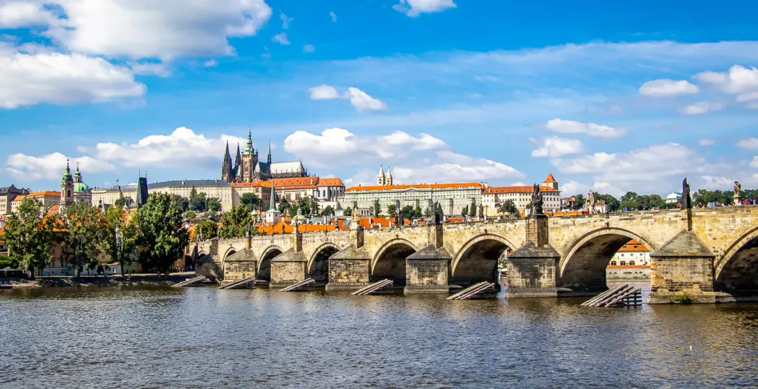 Blick auf die Karlsbrücke mit Statuen und Bögen über einem Fluss, Prager Burg am fernen linken Ufer unter blauem Himmel mit W