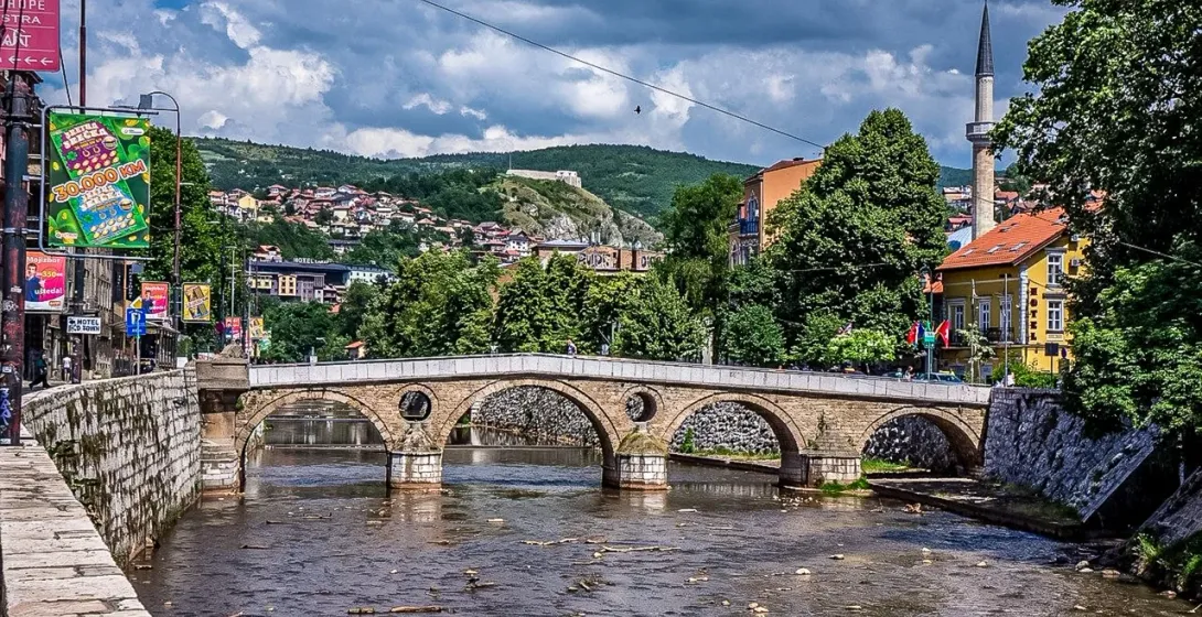 Historische Steinbogenbrücke über einen Fluss in Sarajevo, mit einem Minarett, Gebäuden und grünen Hügeln unter bewölktem Him
