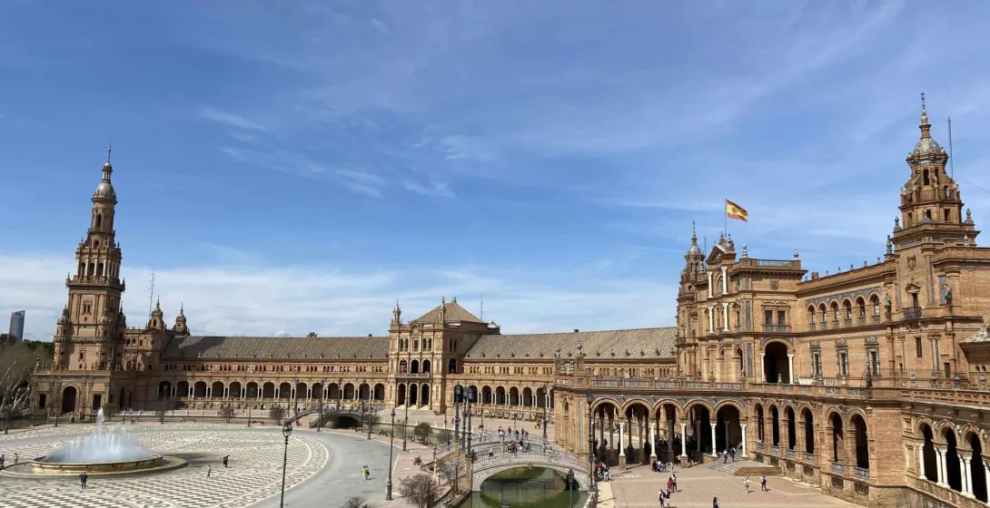 Panoramic view of Plaza de España in Seville, featuring the grand building, canal, bridges, and fountain under a blue sky.