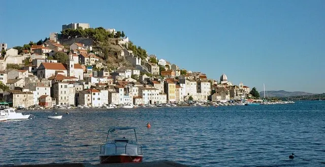 Sibenik old town and hilltop fortress from the sea, showing historic buildings with red roofs, boats, and a clear blue sky.