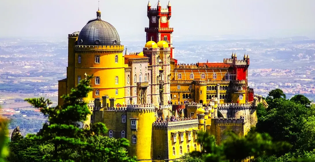 Colorful Pena Palace in Sintra, Portugal, featuring yellow, red, and grey sections with domes and towers, seen from afar thro