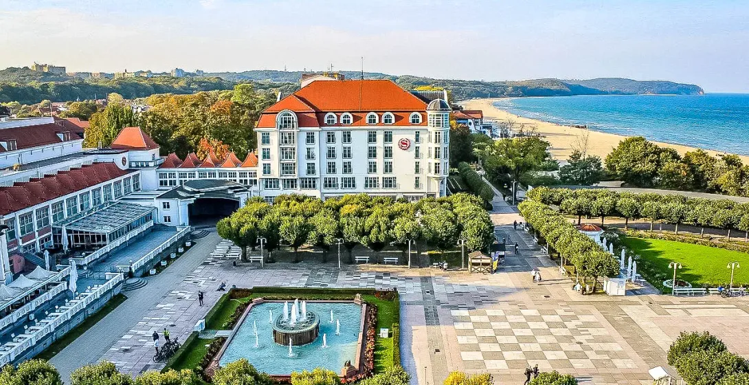 Aerial view of Grand Hotel Sopot with a square, fountain, green trees, sandy beach, and Baltic Sea under a blue sky.