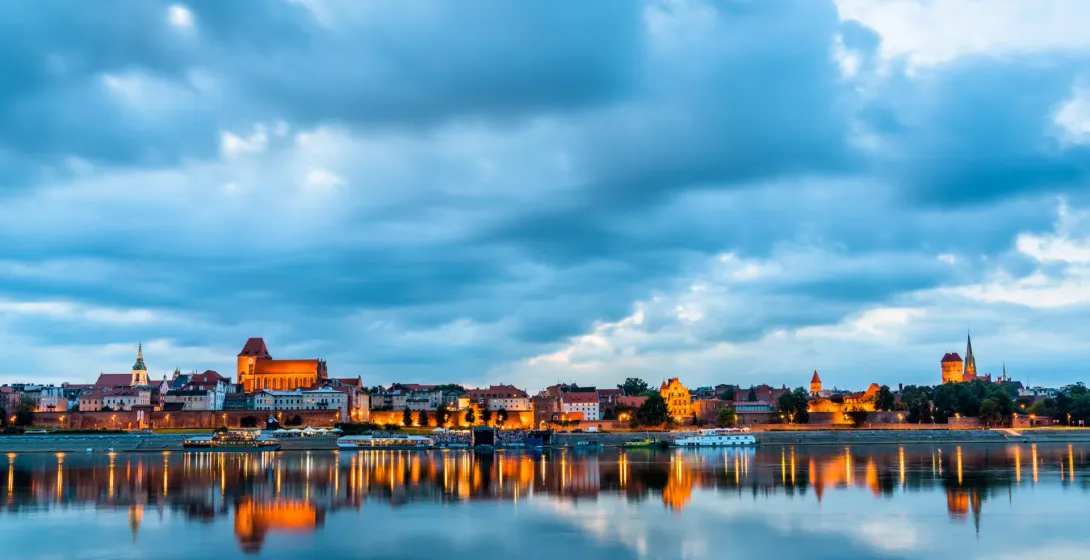 Toruń skyline at dusk, with historical buildings illuminated along the riverbank, reflected in the calm water under a cloudy