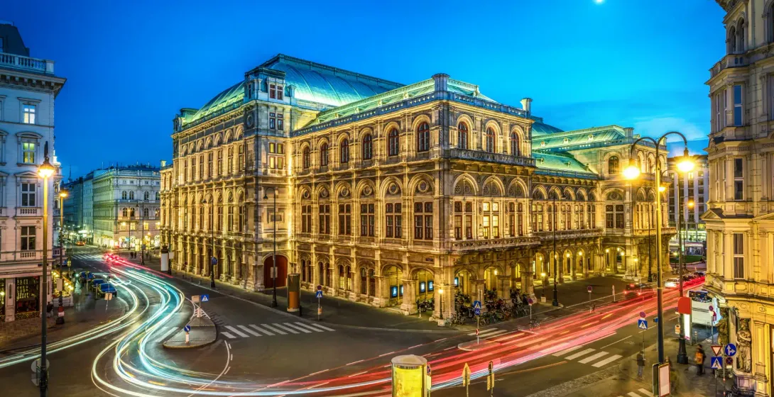 Vienna State Opera at twilight with light trails from cars on the street below and a crescent moon.