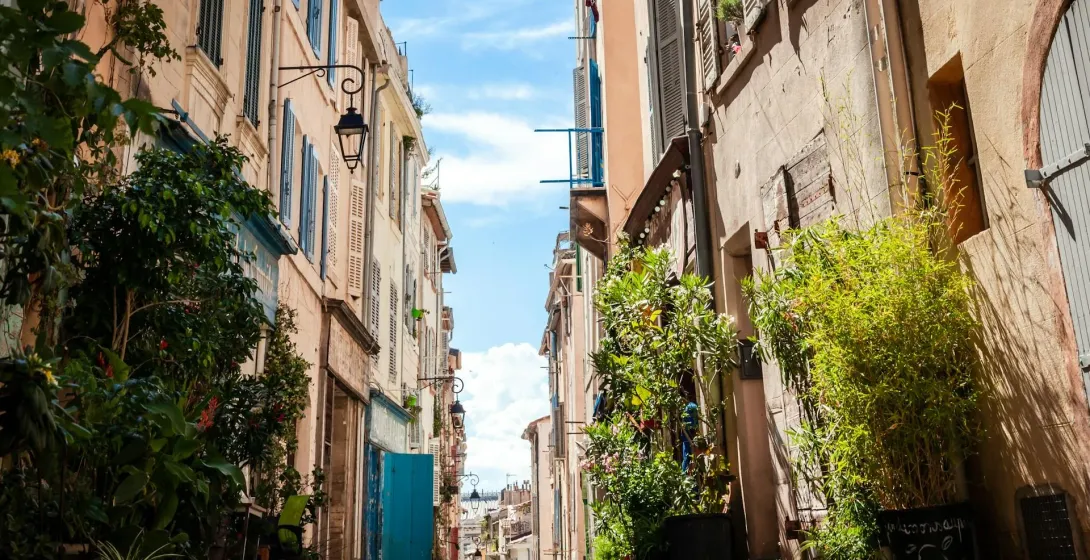Estrecho callejón adoquinado en Marsella con altos edificios antiguos, persianas cerradas y plantas en macetas, bajo un cielo