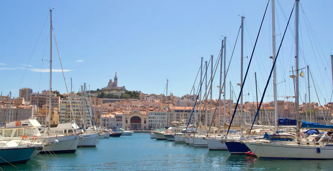 Veleros y yates amarrados en un puerto azul, con el horizonte de la ciudad y la basílica Notre-Dame de la Garde en una colina