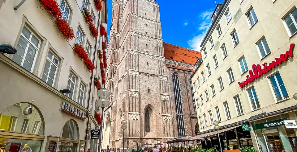 Fußgängerzone in München mit der hohen, zwiebelturmgekrönten Frauenkirche im Zentrum, von Gebäuden umrahmt.