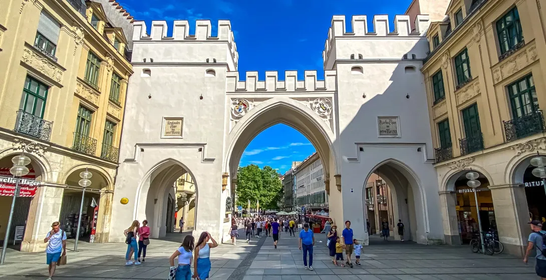 Das Karlstor in München, ein weißes Steintor mit zwei zinnenbewehrten Türmen und drei Durchgängen, bei Sonnenschein mit Passa