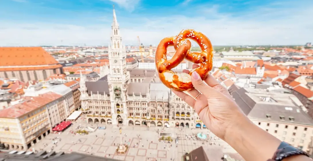 Hand hält eine Brezel mit dem Neuen Rathaus und dem Marienplatz in München im Hintergrund, von oben gesehen.