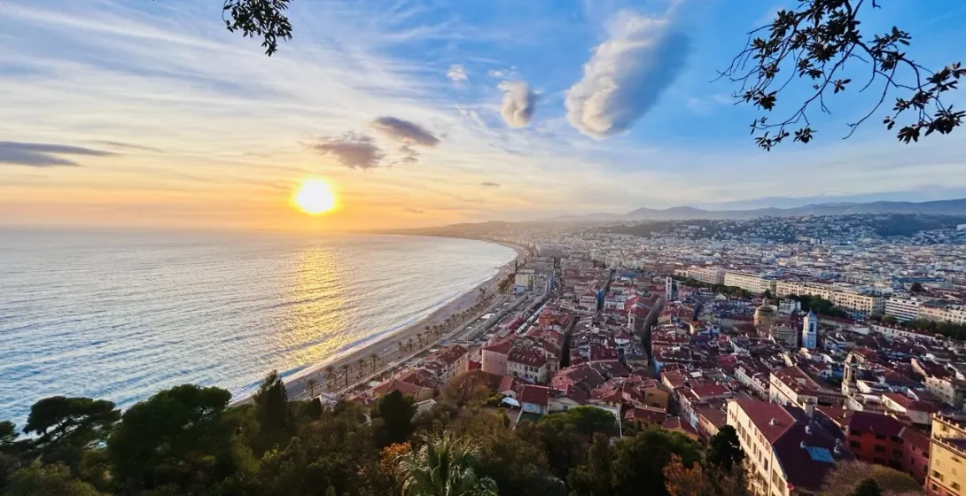 Panoramic view of Nice, France at sunset, showing the curved coastline, city buildings, and a bright sun over the sea.