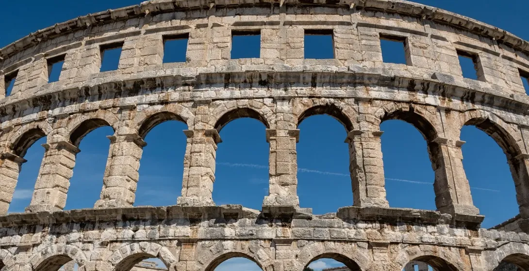 Exterior of the Pula Arena, a Roman amphitheater, with its preserved stone arches against a clear blue sky.