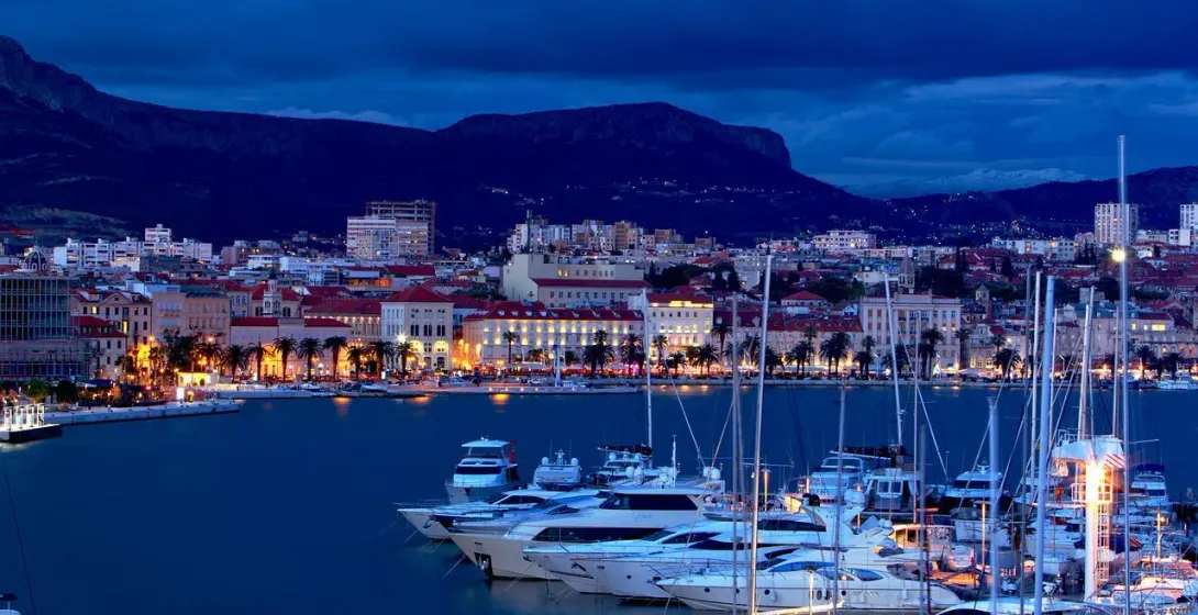 Split harbor at night, with illuminated city buildings, palm trees, mountains, and many yachts.